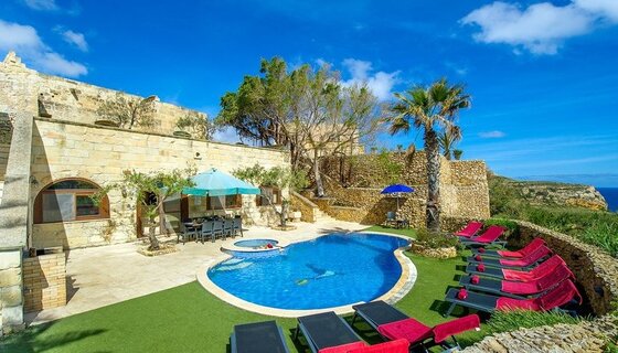 6 bedroom holiday villas in Gozo - Red and black sun loungers face a generous pool in front of a stone building. (© Villa Soldanis)