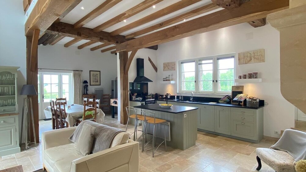 Cuisine Le Figuier Living à Hautefort, France – Poutres apparentes, cuisine avec îlot central, plan de travail en granit noir et sol en travertin. Canapé, tabourets de bar et îlot central en cuir (© Voila Villas France)