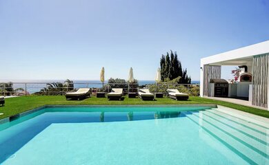 Sun-drenched pool and terrace - A pristine swimming pool with internal steps sits next to a green lawn lined with sun loungers and parasols. In the background, there is a covered outdoor kitchen area with a grill and sea views. (© Goto Property Management)