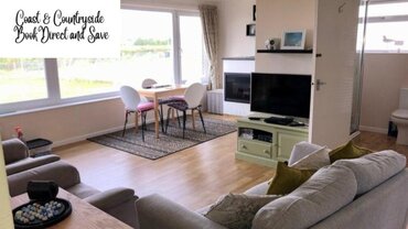 Living room in one of the Norfolk Coast and Countryside properties - Open living room with grey sofa, dining table and chairs and large windows with a view over the green land. Living room in one of the Norfolk Coast and Countryside properties - Open living room with grey sofa, dining table and chairs and large windows with a view over the green land.