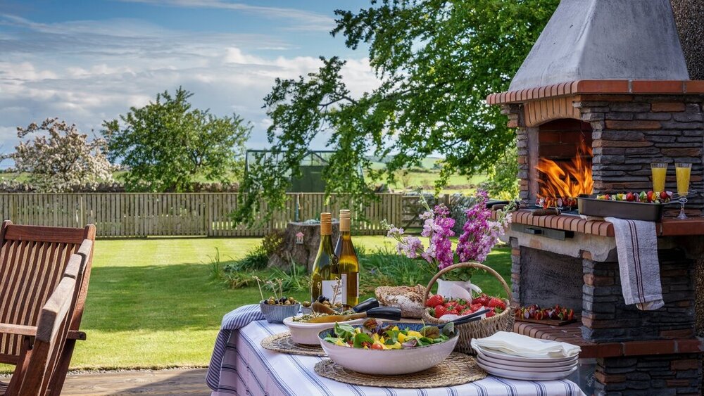 Luxury accommodation in Scotland for intimate birthday gatherings - An outdoor scene with a table holding in wine bottles and plates of food, a wood burner and seating.
