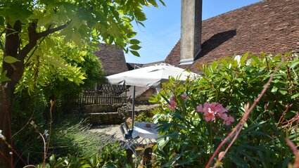 Coin repas intime dans le jardin – Une table dressée pour les repas se trouve sous un grand parasol, dans un espace paisible du jardin. L'extérieur est entouré d'une végétation luxuriante et de murs en pierre, avec le toit de tuiles de la propriété visible en arrière-plan. (© Voila Villas France)