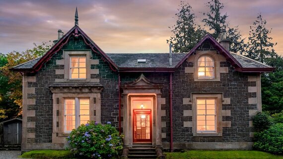 Stay on the banks of Loch Long - A stone building lit from inside in front of a sunset. (© Lochside House)