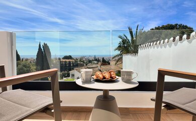 Scenic Balcony Breakfast View - A small table set with two mugs and a plate of croissants sits on a balcony with a glass railing, overlooking a coastal town and distant sea. (© Goto Property Management)