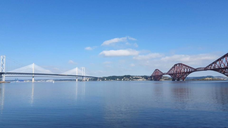 Discover the romance of the South Queensferry bridges - A red rail bridge and two white road bridges cross a wide river.