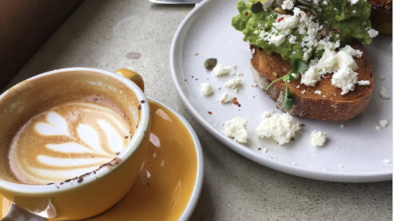 Coffee and - A yellow coffee mug with a creamy coffee alongside a plate with toast, avocado and cream cheese.