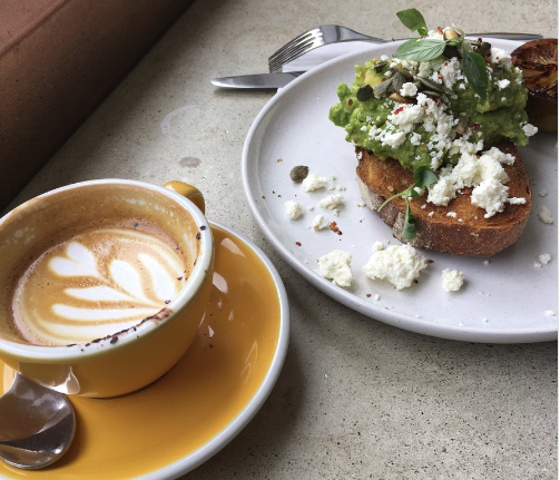 Coffee and - A yellow coffee mug with a creamy coffee alongside a plate with toast, avocado and cream cheese.