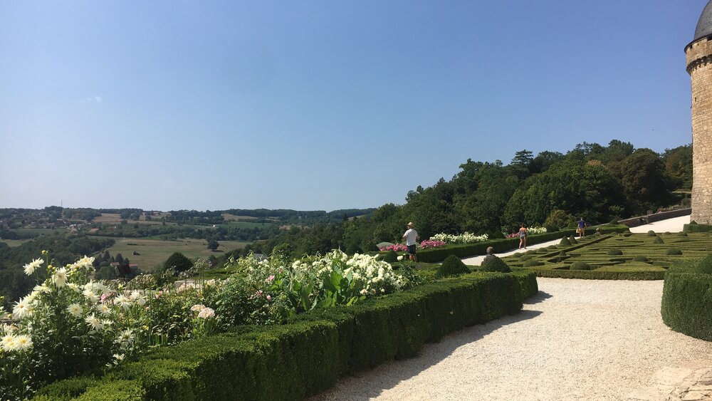 Jardins du château de Hautefort – Haie topiaire basse bordée de fleurs blanches. Ciel d'été bleu et chaud avec une allée sinueuse dans les jardins du château (© Voila Villas France)