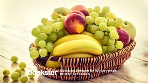 Provide seasonal fruit in a welcome pack - A photo of grapes, bananas and peaches in a wicker fruit basket with a Bookster logo.