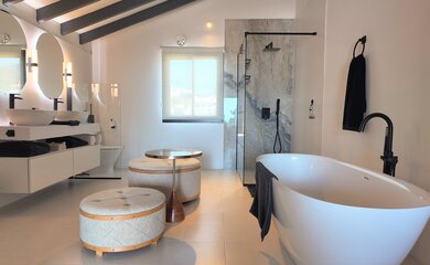 Modern luxury bathroom with tub - A spacious, contemporary bathroom featuring a freestanding white tub, dual vanity sinks, and a large marble-tiled walk-in shower. The room includes two tufted fabric stools and a small side table in the center. (© Goto Property Management)