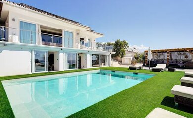 Luxury Villa with Swimming Pool - A modern two-story villa with a turquoise swimming pool, surrounded by artificial grass and several sun loungers. A shaded outdoor seating area is visible in the background under a wooden pergola. (© Goto Property Management)